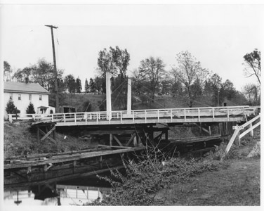 This photograph provides a detailed view of the design of the King-Post style swing bridge that supplanted the A-Frame bridges along the canal.  Originally, A-Frame swing bridges were constructed over the canal crossings.  However, as automobiles and trucks supplanted horse-drawn wagons, the A-Frame bridges were replaced by the stronger King-Post design.  To operate the span, the bridgetender pushed a 10-foot pole and “walked” the bridge, which was balanced on three sets of iron wheels.  The wheels rolled on a circular rail, allowing the bridge to swing to one side of the canal on a turntable.  (Photograph reproduced from the Collection of the Library of Congress)